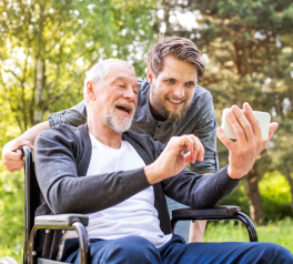 older man in wheelchair taking selfie with young adult standing behind him
